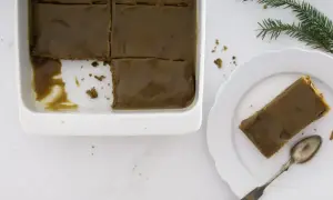 En bit av en saftig sticky toffee pudding med kolaglasyr på en vit tallrik, bredvid en form med resten av kakan.