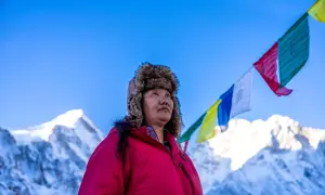 Lhakpa Sherpa, eine Frau mit einem Pelzmützen, blickt in den Himmel, während Gebetsfahnen über schneebedeckten Bergen wehen.