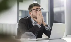 Man med glasögon sitter vid en laptop, gnuggar ögonen. Ett klassiskt dilemma exempel på stressad arbetsdag.
