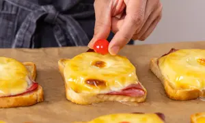 Eine Hand platziert eine Kirsche auf einem überbackenen Toast Hawaii. Der Toast im Backofen ist mit Schinken, Ananas und geschmolzenem Käse belegt.