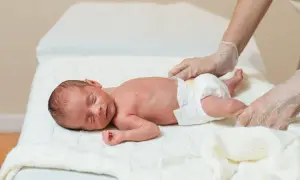 Noworodek z hipotrofią płodu leży na białym kocyku, a ręce w rękawiczkach delikatnie go dotykają.