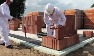Dwóch budowlańców w białych kombinezonach i kaskach układa cegły na fundamencie.