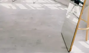 Ett ljust rum med golv i microcement. Solens strålar skapar mönster på golvet. En stor spegel med träram står lutad mot väggen.
