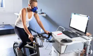 Femme en masque respiratoire effectuant un test d'effort duree sur un vélo d'appartement, connectée à un équipement médical.