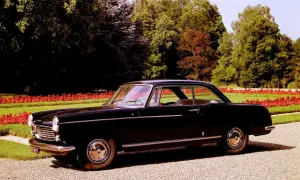 Elegancki, czarny Peugeot 404 coupe z lat 60. stoi na żwirowej alejce, otoczony zielenią i czerwonymi kwiatami.