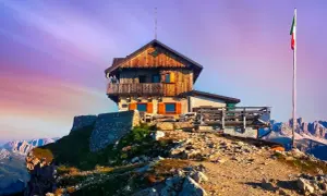 Rifugio Nuvolau thront auf einem Felsvorsprung unter einem lila Himmel. Eine italienische Flagge weht im Wind.