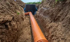 Pomarańczowa rura kanalizacyjna biegnie przez wykopany rów melioracyjny, prowadząc do czarnej studzienki.