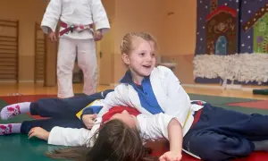 Niñas sonriendo mientras practican judo para niños. Un instructor de judo con cinturón rojo observa.