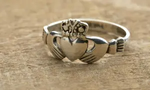 Anillo Claddagh de color oro blanco, con corazón, manos y corona, sobre madera.