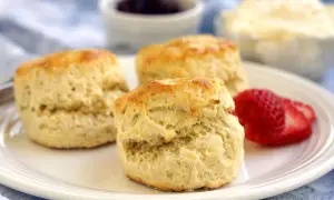 Tre fluffiga scones på en tallrik med jordgubbar, grädde och sylt. Perfekt för fika med de godaste scones.