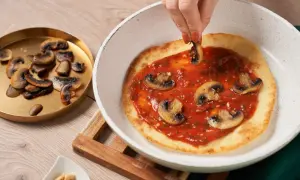 En hand lägger champinjoner på en pizza i stekpanna med tomatsås.
