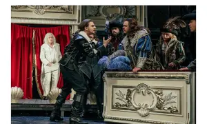 Aktorzy w strojach z epoki na scenie, w tym mężczyzna w czarnym stroju, przypominający Cyrano de Bergerac, gestykuluje do towarzyszy.