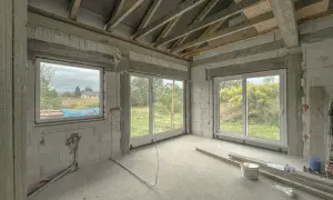 Budowa domu w stanie surowym. Wnętrze z betonową podłogą, ścianami z bloczków i oknami. Widać materiały budowlane, które mogą wpłynąć na tynki cementowo wapienne cena.