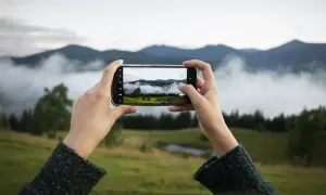 Dłonie trzymają smartfon, na którego ekranie widać krajobraz górski z mgłą i drzewami.