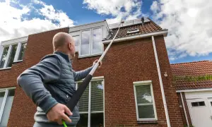 Hombre con pértiga extendida para limpiar tejas de una casa de ladrillo rojo bajo un cielo azul con nubes.