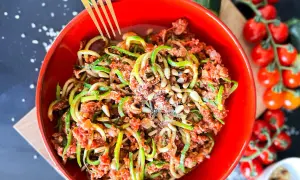 Ein leckeres Zoodles Rezept mit Hackfleischsauce, Pinienkernen und Parmesan in einer roten Schüssel. Daneben Kirschtomaten und Zucchini.