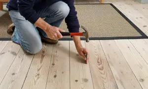 Personen knäböjer och använder en hammare för att montera golvspånskiva på ett trägolv.