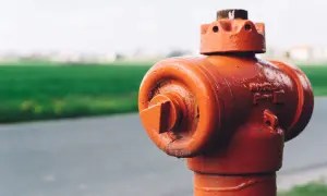 Pomarańczowy hydrant stoi blisko drogi, a za nim widać zielone pole. Odległość hydrantu od ogrodzenia jest niewielka.