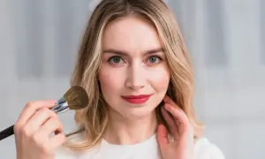 Kobieta z blond włosami i zielonymi oczami nakłada puder pędzlem. Jej usta są pomalowane na czerwono.