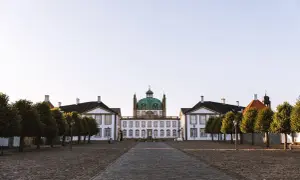 Frederiksborgs slott med sin gröna kupol reser sig majestätiskt mot en klar himmel. En stenlagd väg leder fram till den imponerande byggnaden, flankerad av träd.