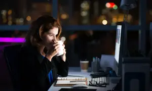 Kvinna dricker kaffe vid sitt skrivbord sent på kvällen, med stadens ljus i bakgrunden. Hon verkar jobba över, kanske för att ta igen komp tid.
