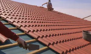 Un tejado en proceso de colocación de tejas rojas. Un trabajador con guantes usa una paleta, mientras otro se sienta en la cima.
