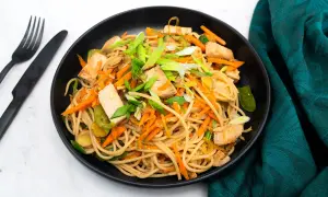 Pyszne makaron z tofu, marchewką i szczypiorkiem w czarnej misce. Obok czarne sztućce i zielony materiał.