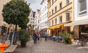 Kempten Geheimtipps: Menschen schlendern durch eine charmante Gasse mit Restaurants und Cafés, umgeben von historischen Gebäuden und üppigem Grün.