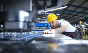 Uśmiechnięty operator CNC w żółtym kasku pochyla się nad maszyną, kontrolując proces produkcji.