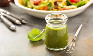 Grünes Dressing in einem Glas mit Salat und Kräutern im Hintergrund.