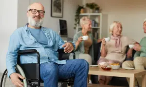 Uomo sorridente in carrozzina, con amici in sottofondo. La sua storia di invalidità civile patologie riconosciute è un esempio di resilienza.