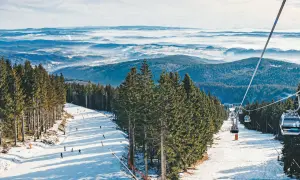 Skifahrer genießen den Winter in Tschechien. Eine Seilbahn schwebt über schneebedeckte Hänge, umgeben von dichten Fichtenwäldern und einer nebligen Berglandschaft.