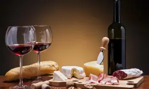 Deux verres de vin rouge, une bouteille, du pain, des fromages, de la charcuterie et des noix. Un accord parfait pour un vin pour fromage et charcuterie.