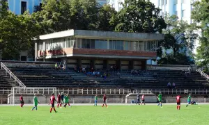 Młodzi piłkarze trenują na boisku. W tle trybuny i budynek, przypominający stadion Dynamo Kijów.