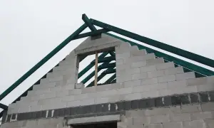 Budowa domu w toku. Widać szczytową ścianę z bloczków, na której powstaje wieniec, oraz zieloną więźbę dachową.
