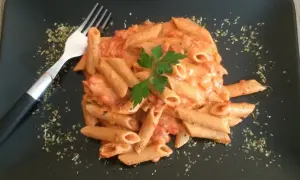 Penne w kremowym sosie pomidorowym z mascarpone, udekorowane natką pietruszki. Obok widelec i posypka z ziół.