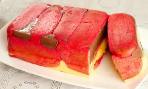 Una fetta di zuppa inglese tradizionale, con strati di pan di Spagna rosa, crema pasticcera gialla e cioccolato, decorata con savoiardi rossi e gocce di cioccolato.