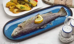 Ein ganzer, im Ofen gebackener Fisch auf einem blauen Teller, garniert mit einer Zitronenspalte. Daneben eine Portion Kartoffel-Gemüse-Spalten.