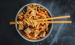 Miska z makaronem udon, tofu i warzywami, przykryta pałeczkami.