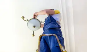 Mężczyzna w niebieskich ogrodniczkach montuje żyrandol na suficie.