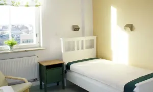 Ett ljust rum med en vit säng, grön sänggavel och en grön nattduksbord. Fönstret visar en stadsvy. Känns som ett rum där någon är inlagd på psyk.