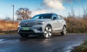 Szary Volvo C40 na mokrej drodze, otoczony suchą trawą i drzewami.
