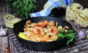 Spaghetti mit Garnelen mediterran in einer Gusseisenpfanne, garniert mit Zitrone und Petersilie. Daneben rohe Pasta, Knoblauch und Chili.