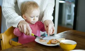 Mama pomaga dziecku kroić rybę na talerzu. To idealne pancakes dla niemowlaka, które uczy się jeść samodzielnie.