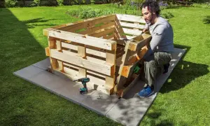 Mann baut ein schmales Hochbeet aus Paletten im Garten. Werkzeug und Schrauben liegen bereit.