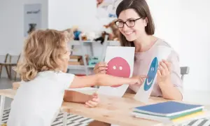 Nauczycielka z uśmiechem pokazuje dziecku karty z buźkami, wspierając jego przygotowanie pedagogiczne.