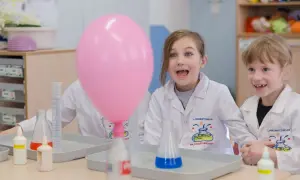 Dzieci w fartuchach laboratoryjnych z ekscytacją obserwują różowy balon podczas eksperymentów chemicznych dla dzieci.