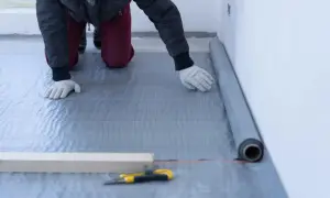Pracownik układa folię pod styropian na piętrze. Materiał izolacyjny jest rozwijany przy ścianie.