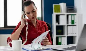 Kobieta w czerwonej bluzce prowadzi rozmowę telefoniczną, przeglądając dokumenty. Może to być element jej pracy związanej z twardą windykacją.