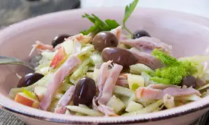 Frischer Fenchel Salat mit Apfelstücken, Schinkenstreifen und dunklen Oliven in einer rosa Schüssel.